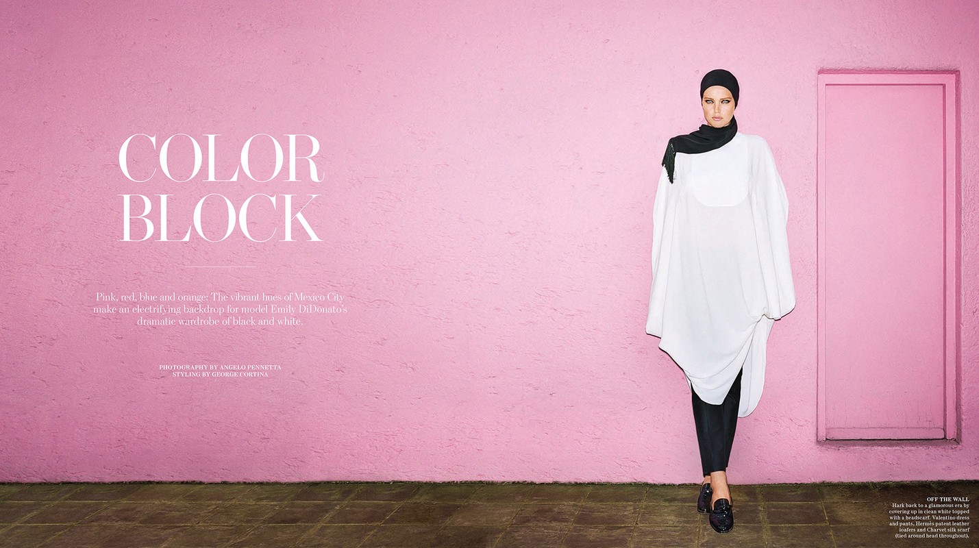 Model Emily DiDonato wearing a white tunic and black headscarf standing against a vibrant textured pink wall.