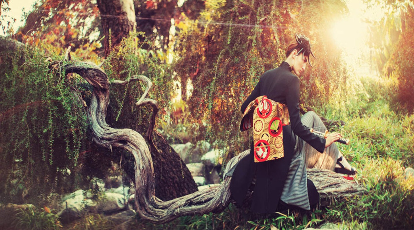Elegant female in kimono sits outdoors near a tree with a sword, sunlight filtering through branches creating a serene and artistic scene.
