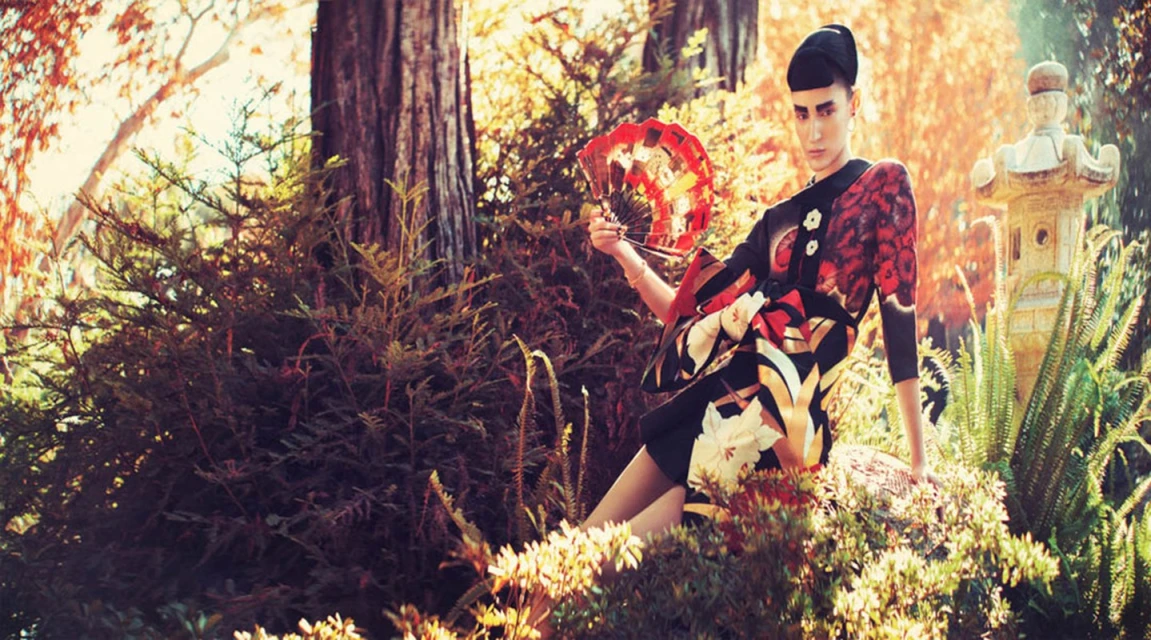 Woman in an ornate black kimono sits amongst greenery and a stone pagoda holding a handheld fan, in a natural setting with filtered light.