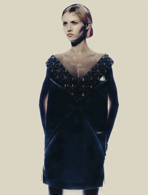 A fashion image featuring a slender adult female model in a dark, V-neck dress with decorative beading, against a soft beige background.