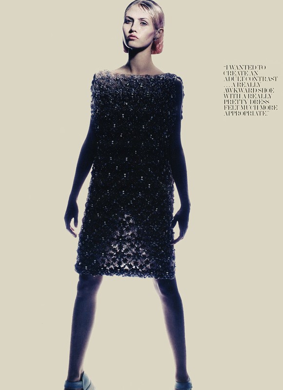 A model in a beaded mini dress stands against a pale backdrop. Full-length shot, studio lighting. Fashion editorial style, neutral colour palette.