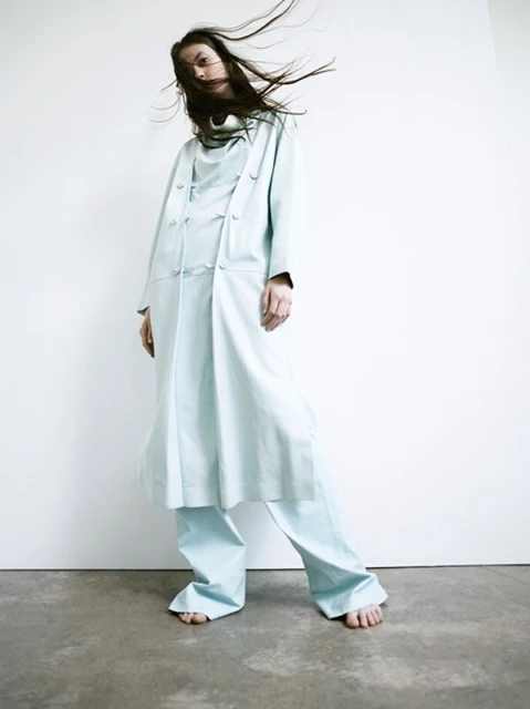Fashion shot of a young adult female wearing a light blue, multi-piece, oversized fashion outfit on a concrete floor in a white studio.