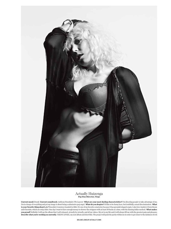 Black and white portrait of a woman with curly blonde hair wearing a bra and robe, posing indoors against a white background.