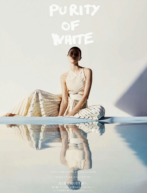 Elegant woman in white outfit sitting near water with reflection, fashion editorial shot, purity of white, studio setting, bright lighting.