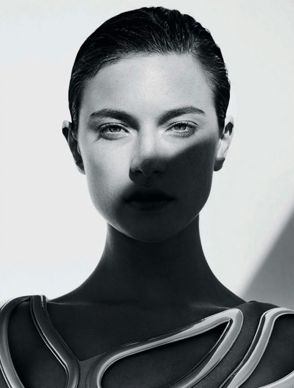 Black and white headshot of a female model with short, slicked-back hair and dark eyes, wearing a futuristic top, against a white backdrop.