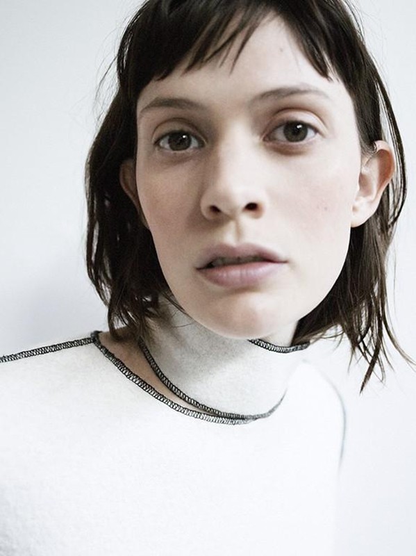 Close-up portrait of a woman with dark short hair and bangs wearing a white turtleneck top with contrast stitching against a white wall.