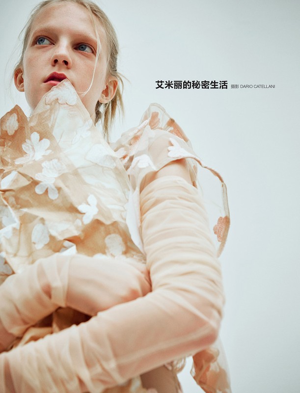 A fair-skinned person with blonde hair poses in a peach-colored floral dress in a light studio environment, looking off-camera.
