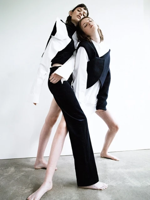 Two fashion models pose together in a photo studio. One model wears black trousers and a black-and-white top. The other model wears a white shirt.