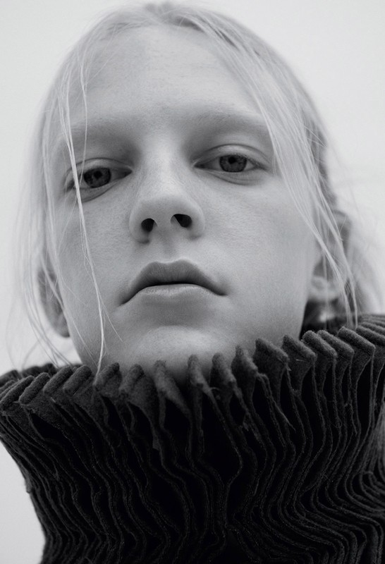 Close-up portrait of a model with blonde hair wearing a high-collared pleated black garment against a plain background.