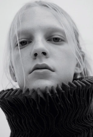 Close-up portrait of a model with blonde hair wearing a high-collared pleated black garment against a plain background.