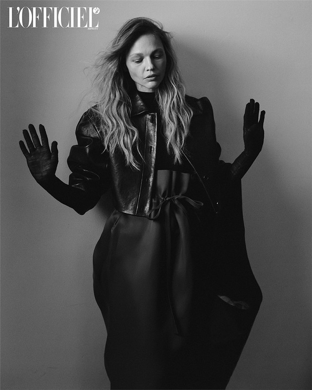 Black-and-white fashion portrait of an adult female model wearing a jacket, gloves, and a flowing dress against a grey background.