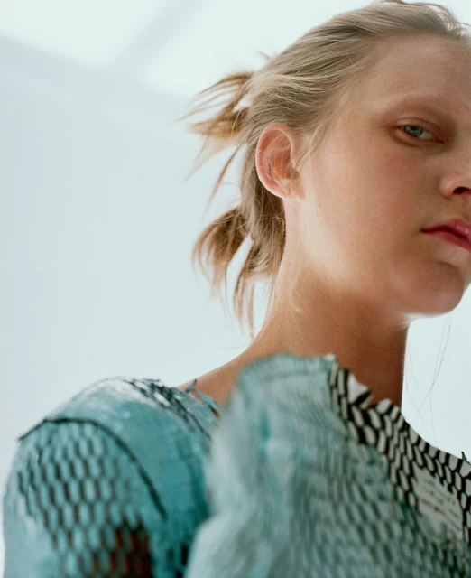 Close-up portrait of a blonde woman with light skin wearing a textured teal and black top against a bright white backdrop in a studio.