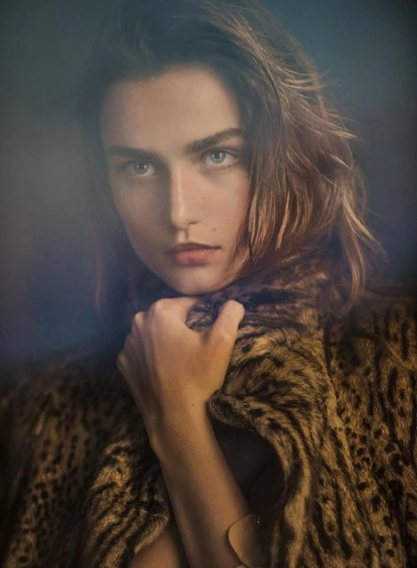 Close-up portrait of a woman with wavy brown hair and light eyes wearing a leopard print coat, gazing directly at the camera.