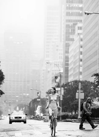 Monochrome street view of a woman riding a bicycle down a wide avenue amidst tall city buildings.