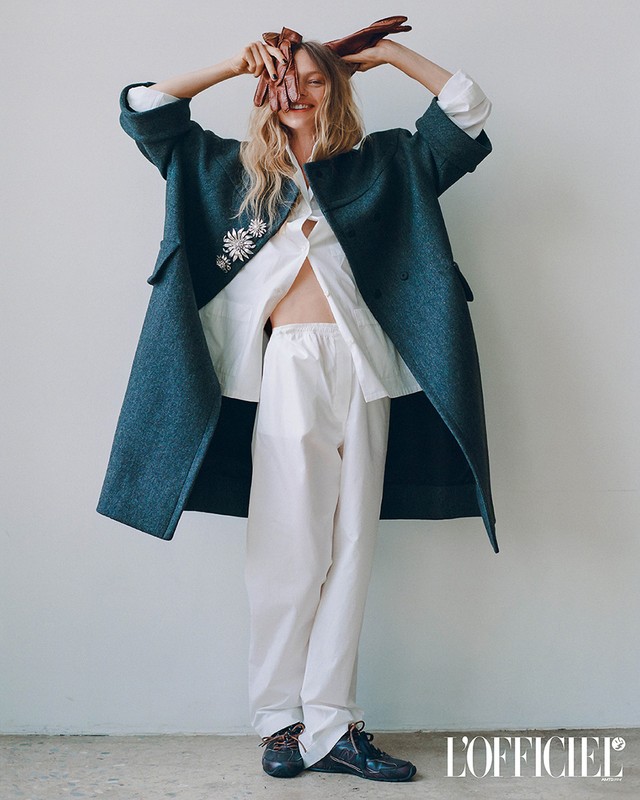 Editorial fashion shot of a woman in a blue coat and white outfit holding gloves, posing against a simple off-white background.