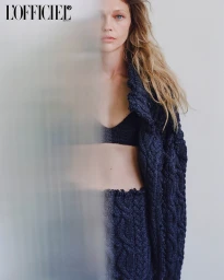 Close-up portrait of a woman with long blonde hair wearing a textured dark blue knitted cardigan against a white background.