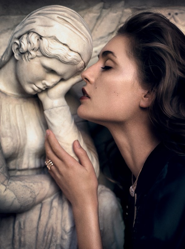 Brunette woman with ring posing with a marble sculpture of a young woman in an indoor environment with moody lighting. 