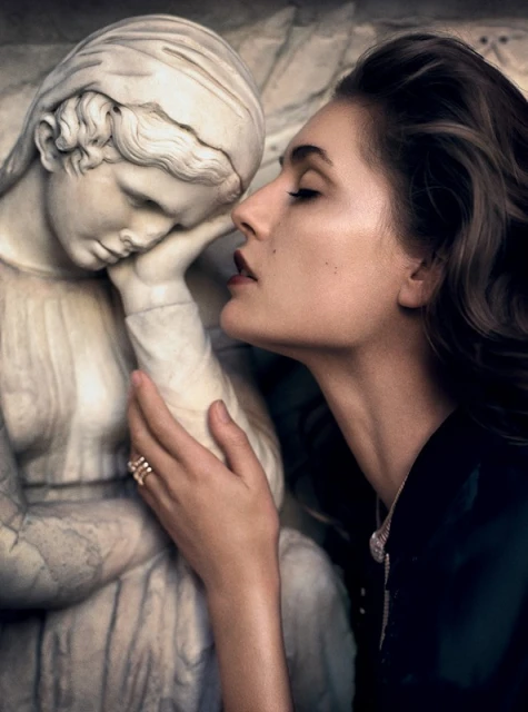 Brunette woman with ring posing with a marble sculpture of a young woman in an indoor environment with moody lighting. 