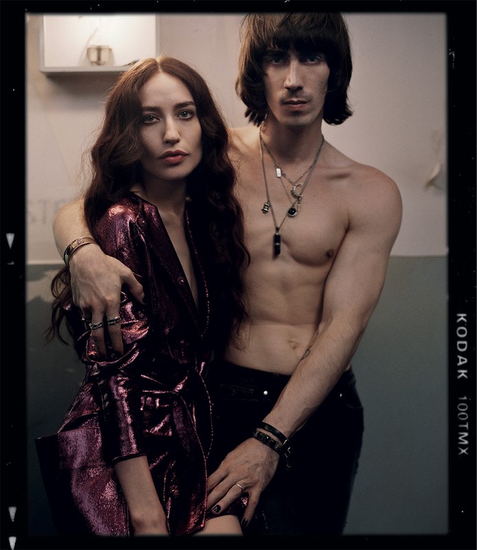 Intimate portrait of a couple, the woman in a shiny purple jacket, the man shirtless with necklaces, in a vintage film border.