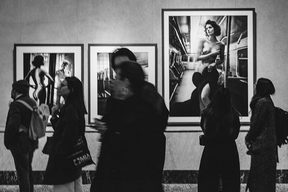 Black-and-white photo of people in an art gallery viewing framed photographs with nude figures inside an indoor setting.