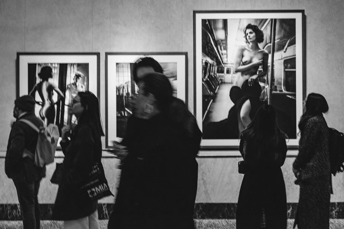 Black-and-white photo of people in an art gallery viewing framed photographs with nude figures inside an indoor setting.