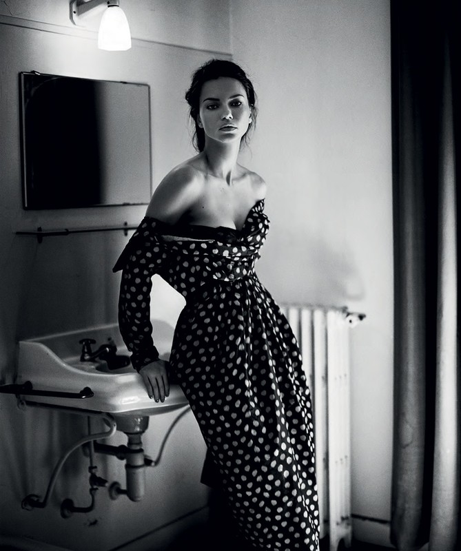 Black and white portrait of a woman in a polka-dot dress leaning against a vintage sink in an indoor bathroom setting with mirror and radiator.