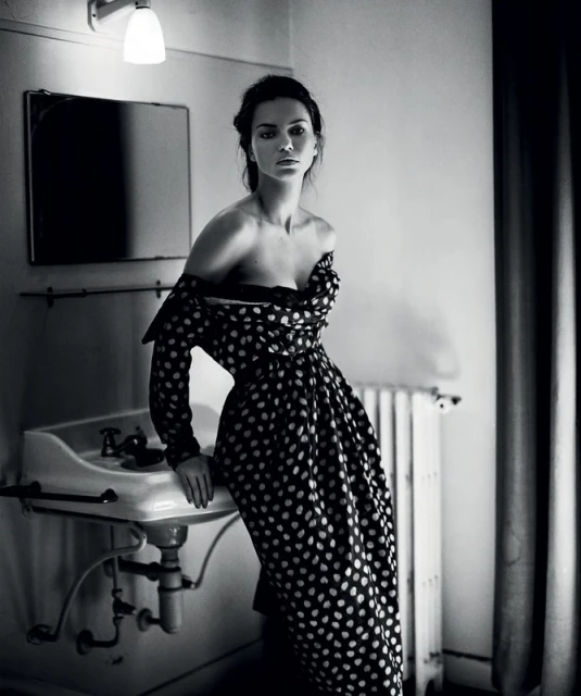 Black and white portrait of a woman in a polka-dot dress leaning against a vintage sink in an indoor bathroom setting with mirror and radiator.