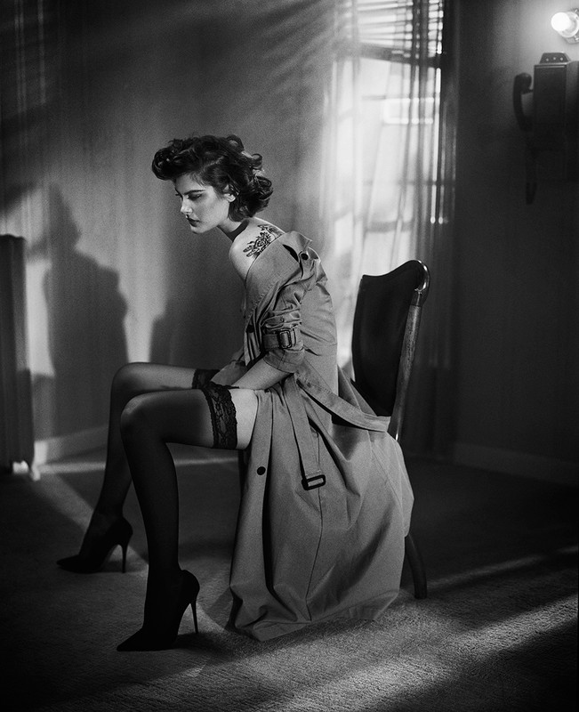 Woman in trench coat and stockings seated on a chair in a moody room by Vincent Peters