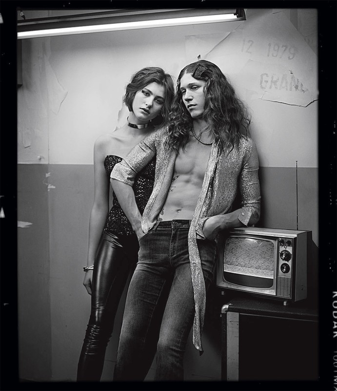 Black and white portrait of a young adult couple in a grungy setting, with the man leaning on an old television set.