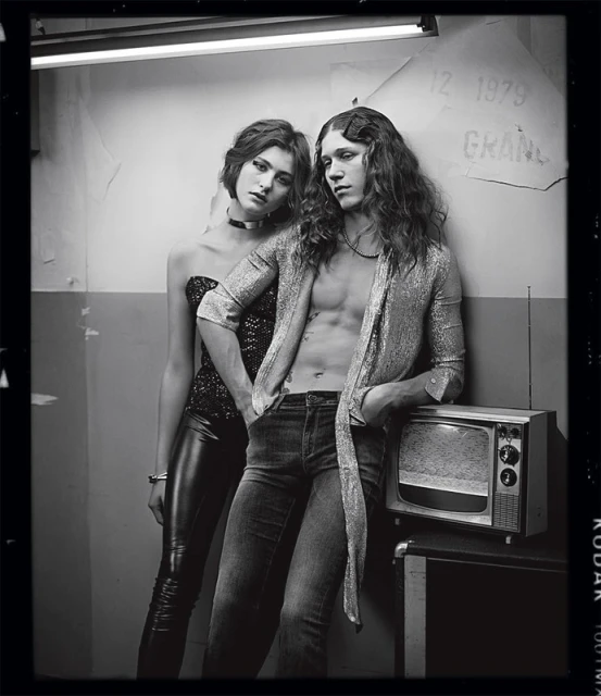 Black and white portrait of a young adult couple in a grungy setting, with the man leaning on an old television set.