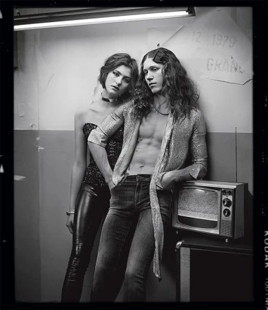 Black and white portrait of a young adult couple in a grungy setting, with the man leaning on an old television set.