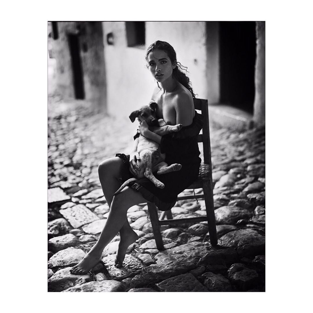 Monochrome portrait of a young adult female sitting on a chair, holding a puppy in her arms, with an outdoor stone street backdrop.