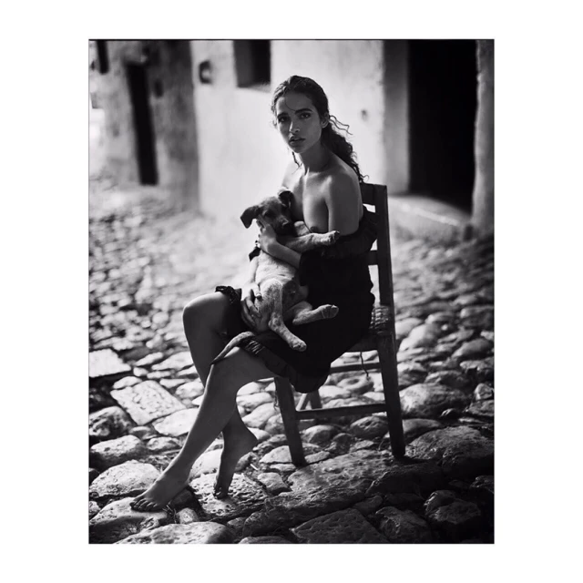 Monochrome portrait of a young adult female sitting on a chair, holding a puppy in her arms, with an outdoor stone street backdrop.