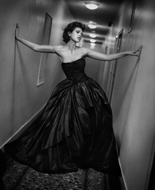 A full-length black and white photo of a female fashion model in a strapless black dress standing in a hallway with her arms extended.