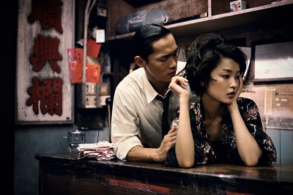 A stylish Asian couple is inside a cafe or restaurant. The man has his hand on the woman's shoulder as she leans on the counter.