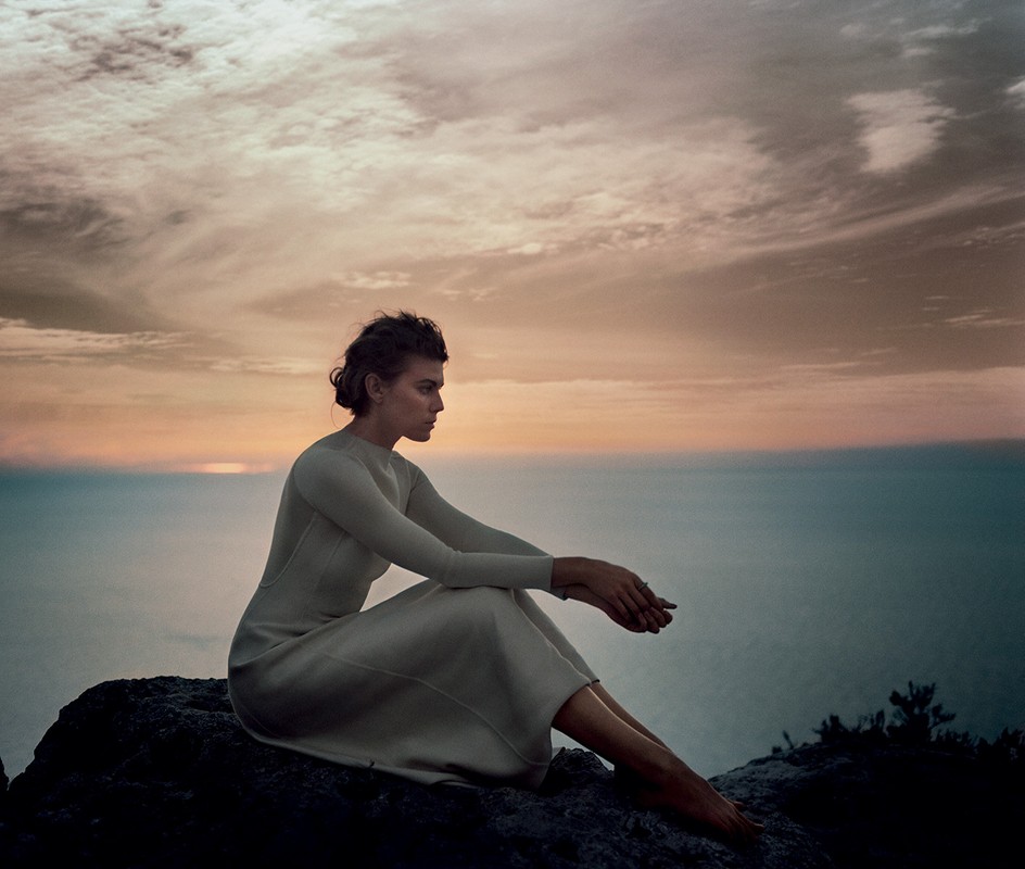 Woman in long-sleeve dress sits on a rock by the ocean at sunset. Dramatic clouds fill the background sky. Soft, warm light.