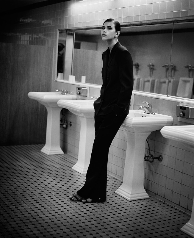 Black and white shot of an adult female in a suit leaning against a white sink in a tiled public restroom with a mirror and bright lighting.