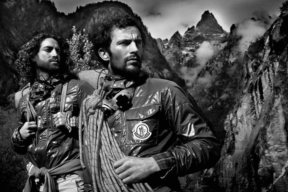 Two men pose in shiny jackets with rope against a mountain backdrop. Monochrome fashion shot with dramatic lighting.