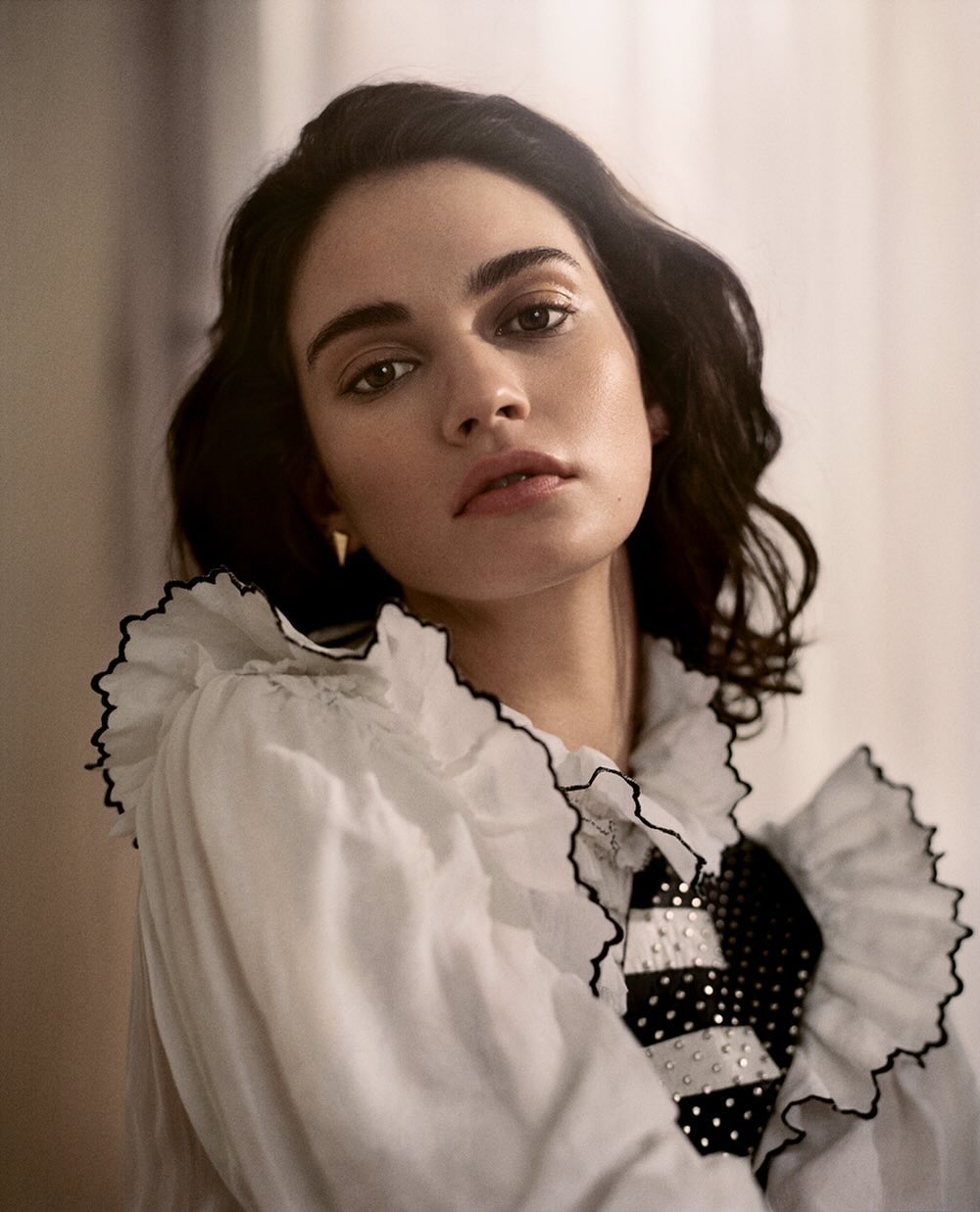 Portrait of a brunette woman wearing a white ruffled shirt with black trim, posing indoors in a brightly lit room.