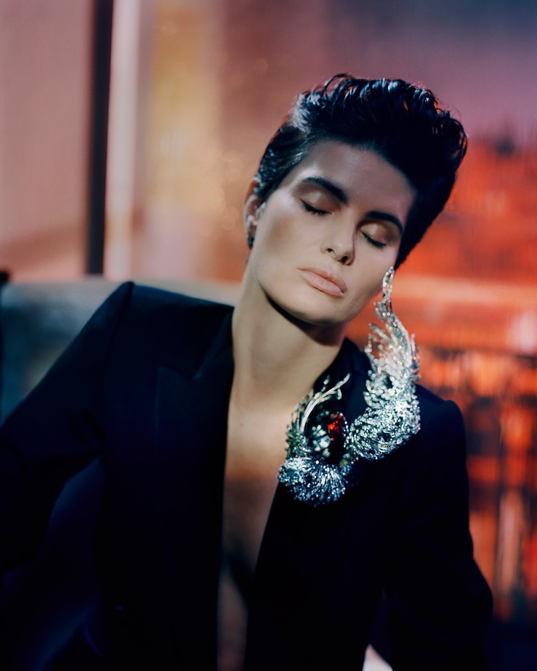 Woman with a low haircut poses wearing a black blazer with a large, ornate brooch. Indoor setting with blurred warm-toned background.