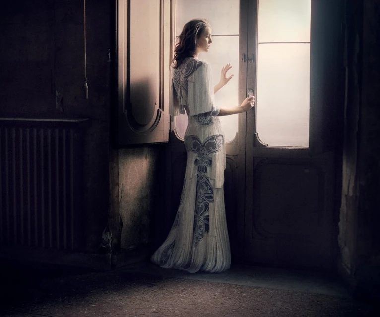 Elegant woman in ornate white dress stands at glass doors in dimly lit room, exuding classic fashion and vintage beauty.