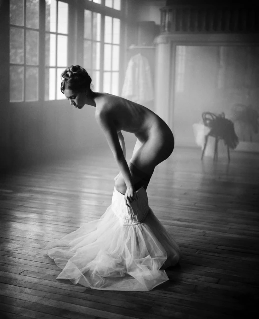 Black and white image of a nude woman putting on a white tutu in a sparse room with wooden floors and large windows.