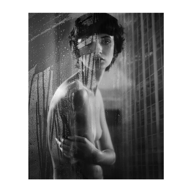 Monochromatic portrait of a topless, dark-haired woman behind frosted glass with visible water droplets, looking directly at the camera.