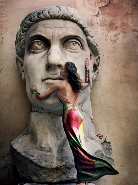 Woman in gown leans against a large stone sculpture head in an outdoor setting with textured walls and artistic lighting.