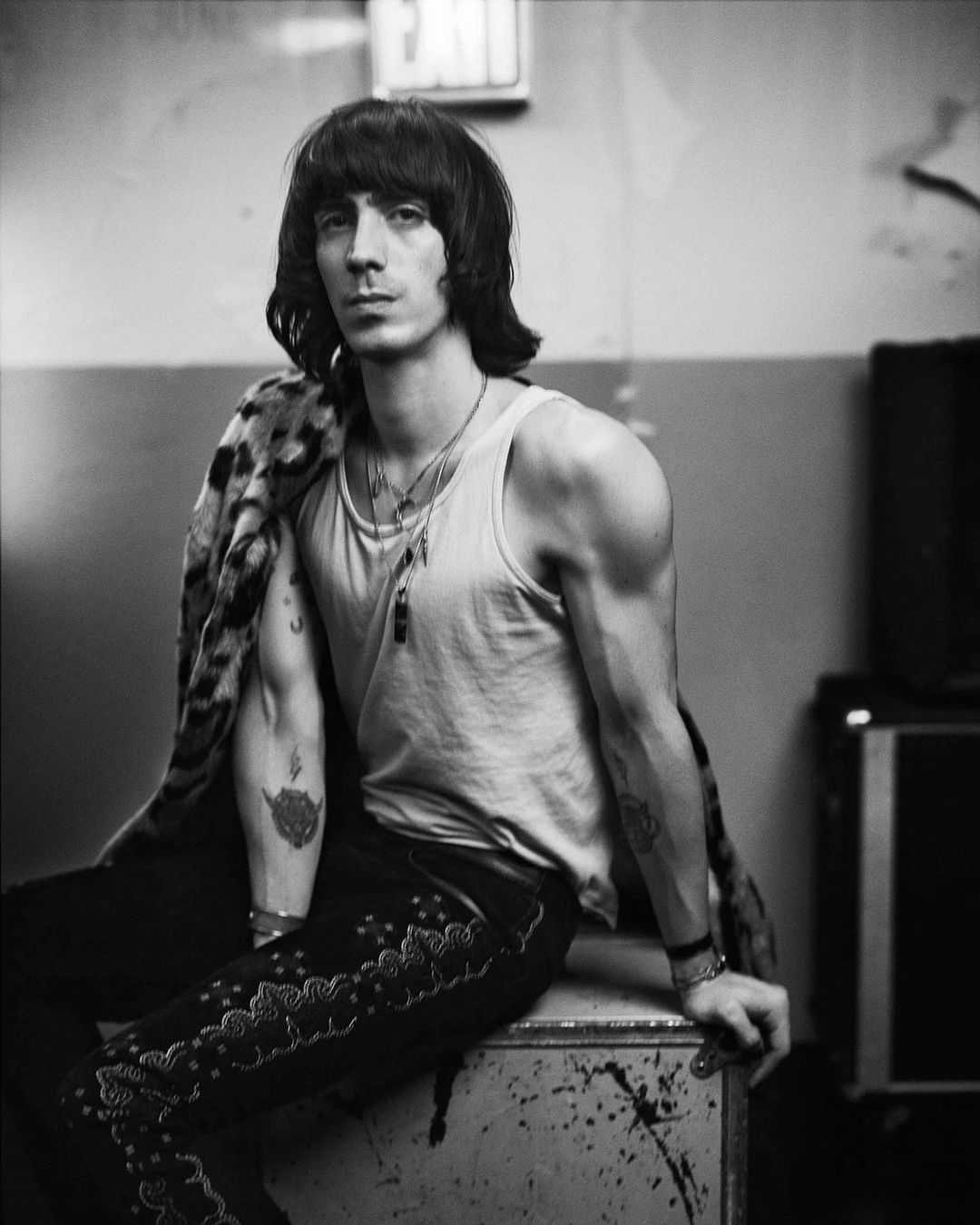 Black and white portrait of a slender adult male with dark hair wearing a tank top and animal print coat, sitting on a metal box.