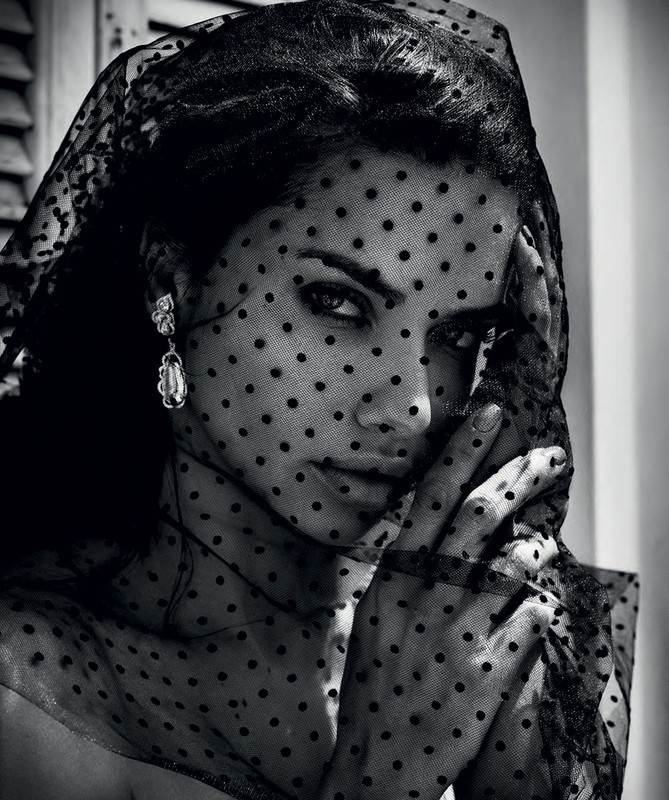 Black and white portrait of a woman wearing a black, polka-dot veil and dangling earrings, with a hand near her face.