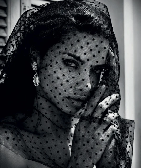 Black and white portrait of a woman wearing a black, polka-dot veil and dangling earrings, with a hand near her face.
