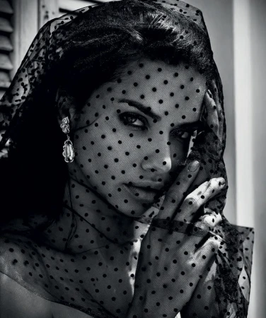 Black and white portrait of a woman wearing a black, polka-dot veil and dangling earrings, with a hand near her face.