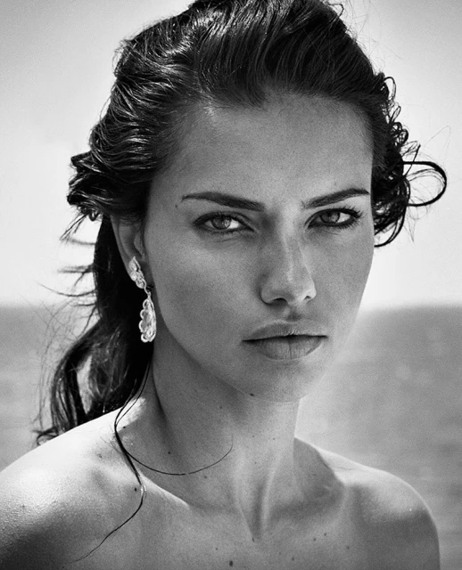 Striking black and white portrait of a woman with wet hair, gazing directly at the camera, wearing dangling earrings against a blurred ocean.