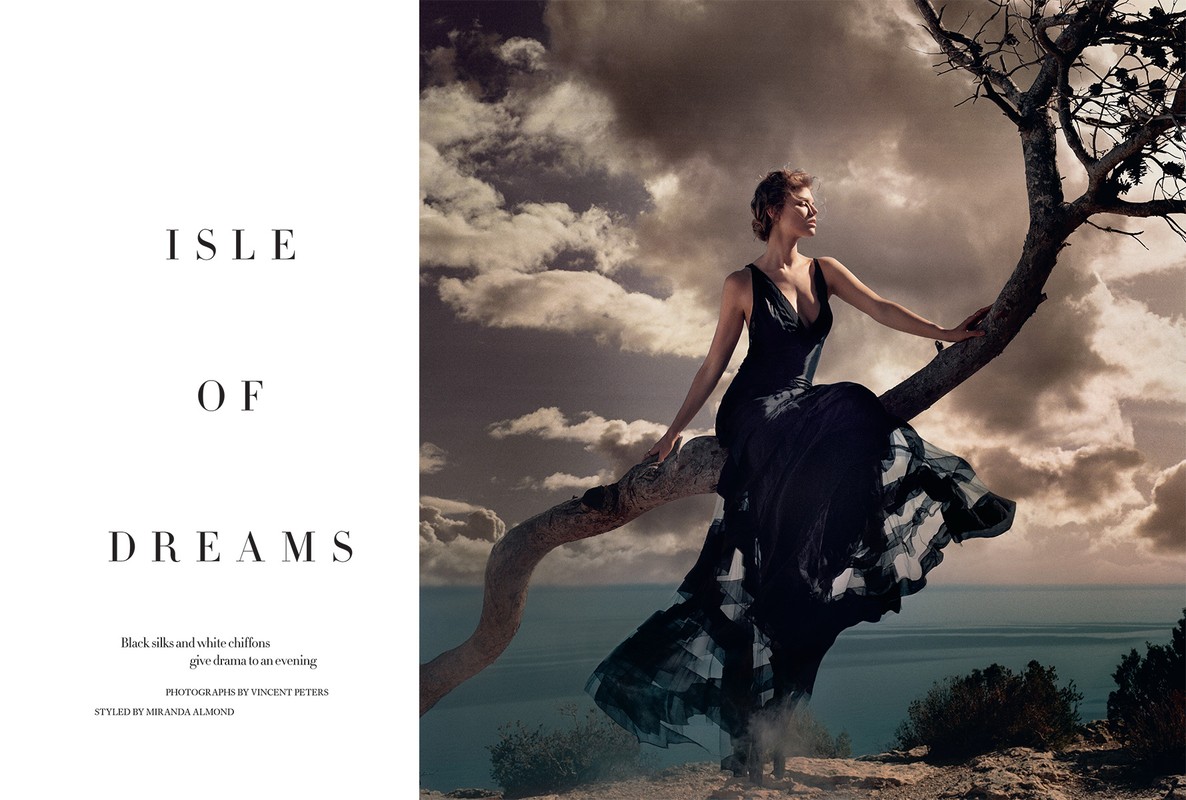 Elegant woman in black dress sits on tree branch against dramatic cloudy sky and ocean backdrop in a fashion editorial style photoshoot.